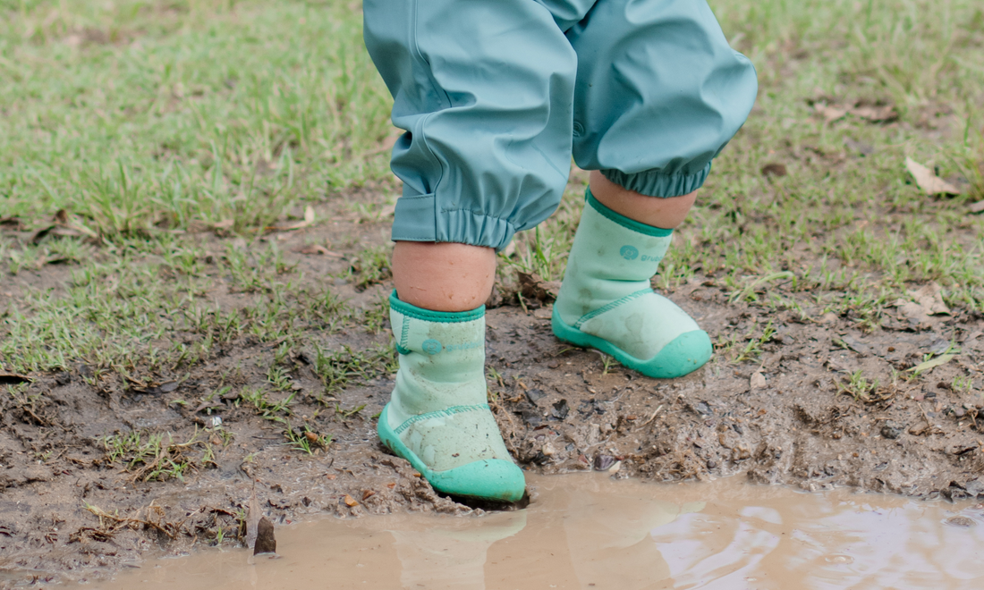 Toddler in green wide toe wee wellies and waterproof rain pants, cautiously stepping into a puddle