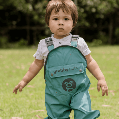 A child wearing green waterproof overalls with adjustable straps standing on grass.