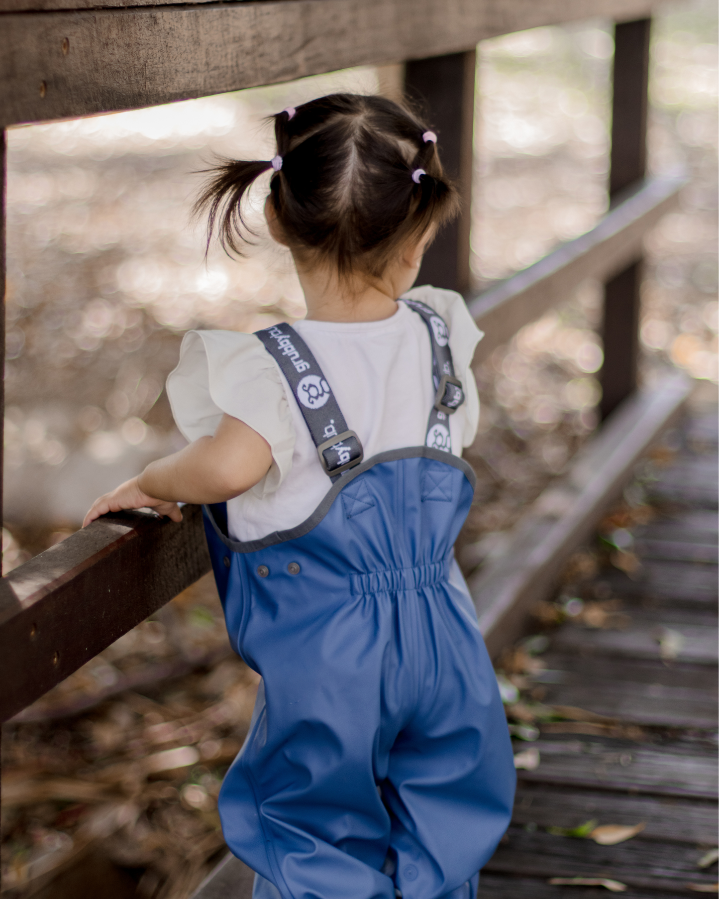 Child in sustainable, waterproof blue overalls standing on a wooden bridge