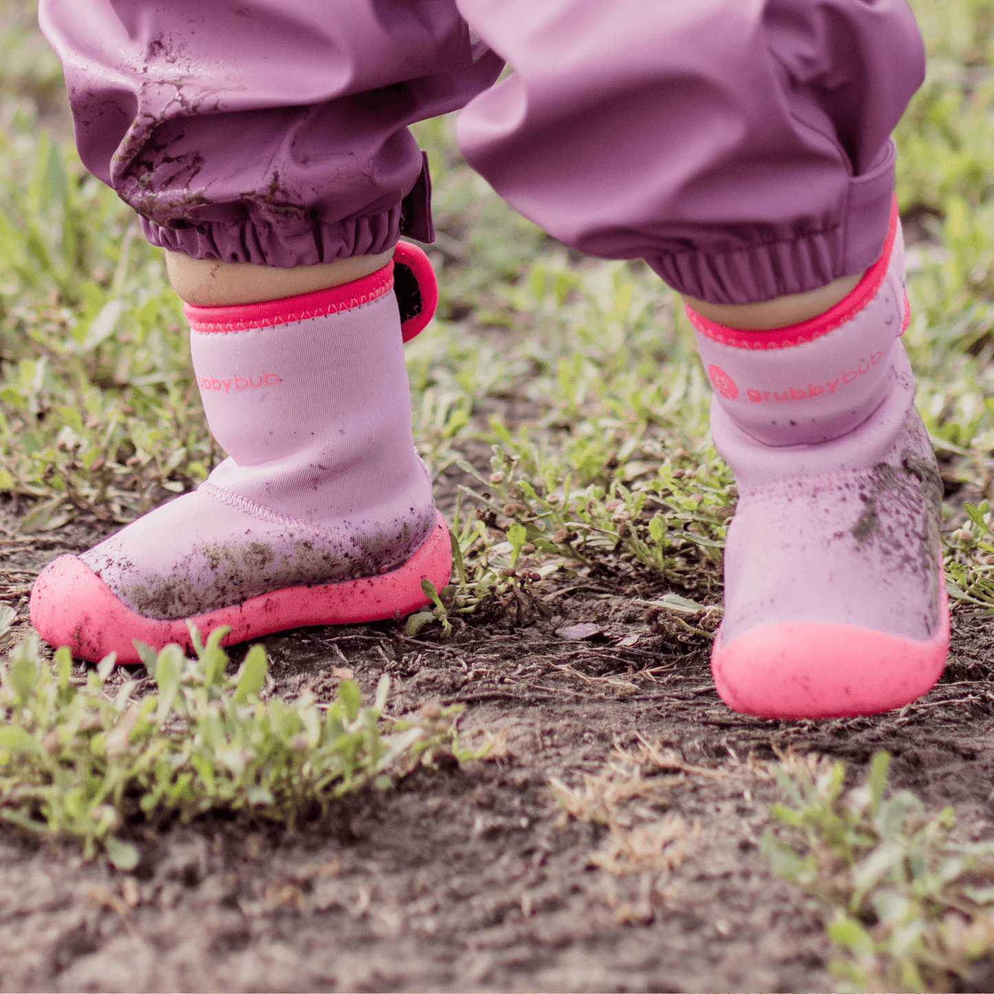 A pair of pink Wee Wellies with an adjustable calf opening, designed for toddlers. The booties are shown with child walking over muddy ground