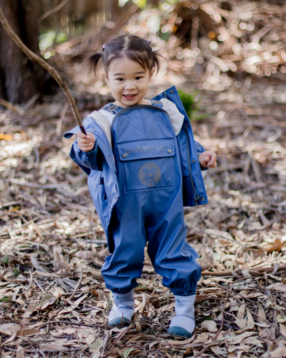 Child in a blue raincoat and waterproof rain pants standing in a forest with dry leaves on the ground.